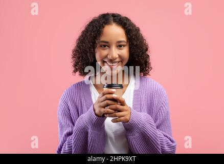 Glückliche junge schwarze Frau, die Kaffee zum Mitnehmen in einer Einweg-Papiertasse hält und auf rosa Hintergrund vor der Kamera lächelt Stockfoto
