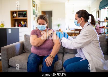 Kaukasische reife Ärztin, die zu Hause eine reife kaukasische Frau mit Covid-19-Impfstoff impft Stockfoto