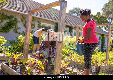 Lächelnde kaukasische reife Frau, die afroamerikanische Tochter anschaut, die Pflanzen wässert, während sie im Garten arbeitet Stockfoto