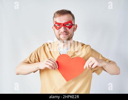 Fröhlich kaukasischen männlichen Kardiologen Arzt in medizinischen Uniform und roten Superhelden Augenmaske mit rotem Papier Herzform. Erwachsene Arzt Chirurg im Krankenhaus posiert über der Wand. Hochwertige Fotos Stockfoto