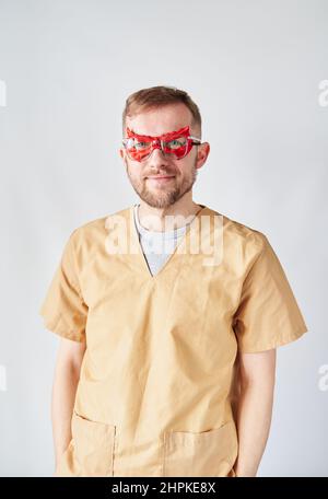 Porträt eines bärtigen Arztes oder einer Krankenschwester mit roter Superhelden-Augenmaske und Brillen und beiger Uniform. Hübscher kaukasischer Arzt, der im Krankenhaus posiert. Hochwertige vertikale Fotos Stockfoto