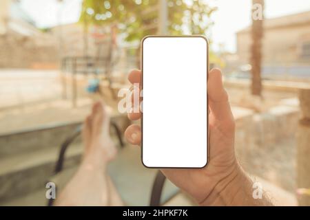 Entspannter Mann, der auf der Terrasse des Apartments sitzt, mit den Füßen auf dem Tisch und Blick auf den Modusbildschirm des Mobiltelefons, selektiver Fokus Stockfoto