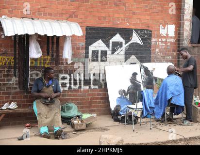 (220222) -- HARARE, 22. Februar 2022 (Xinhua) -- Ein Friseur und ein Schuhreparatureur arbeiten in Mbare, Harare, Simbabwe, 8. Februar 2022. Für viele Besucher scheint Mbare, eine der einkommensschwachen Siedlungen von Harare, eine chaotische und schmutzige Stadt zu sein. Die staubigen Straßen der Gegend, die baufälligen Wohngebäude und die überfüllten Straßenmärkte tun der Siedlung keinen Gefallen. Trotz all des Chaos, das Mbare charakterisiert, zeigt ein genauerer Blick, dass inmitten all dieses Chaos fleißige Bürger ihr Bestes geben, um in einem schwierigen wirtschaftlichen Umfeld ihren Lebensunterhalt zu verdienen. ZUM FEATURE: Histor Stockfoto