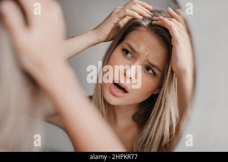 Frustrierte Frau sucht Haarflocken leiden an Schuppen Problem, Blick auf ihre Reflexion im Spiegel im Badezimmer Stockfoto