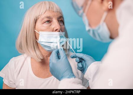 Nahaufnahme eines Arztes in einem medizinischen PSA-Anzug nimmt die Probe von der Nase eines Patienten zum Covid-19-Schnellantigen-Test auf Coronavirus auf blauem Hintergrund Stockfoto