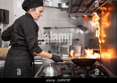 Frau Chefkoch Kochen Wok in der Küche. Kochen flammender Wok mit Gemüse in der kommerziellen Küche. Hochwertige Fotografie. Stockfoto