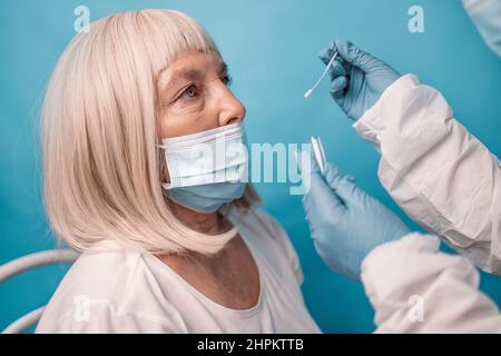 Arzt in medizinischer Maske, die Covid-19 diagnostisches Verfahren für 50s 60s Frauen wegen Coronavirus auf blauem Hintergrund im Labor macht. Stockfoto