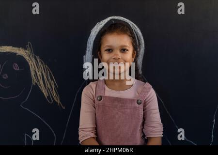 Kleines Mädchen, das gegen eine Tafel steht, mit Zeichnung im Spielzimmer, mit Blick auf die Kamera. Stockfoto