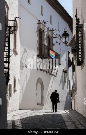 Mann geht auf einer schmalen Straße in Sitges, Spanien. Rückansicht. Sonnenlicht trifft auf die Gebäudefassade. LGBT-Gemeinschaftsflagge hängt auf einer Terrasse. Stockfoto