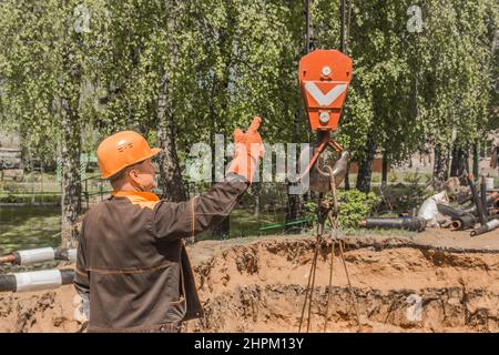 Weißrussland, Region Minsk - 20. Mai 2020: Der Industriearbeiter in den Overalls und dem Schutzhelm nimmt mit seiner Hand den schweren Hakenhebemechanismus des Balkens Stockfoto