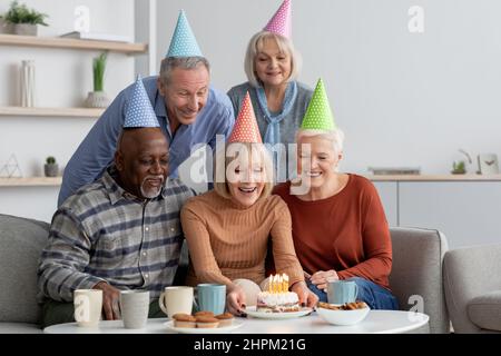 Eine multiethnische Gruppe älterer Menschen, glückliche Männer und Frauen mit bunten Papierhüten, die zu Hause eine Geburtstagsfeier feiern, kaukasische ältere Dame, die eine Geburt hält Stockfoto