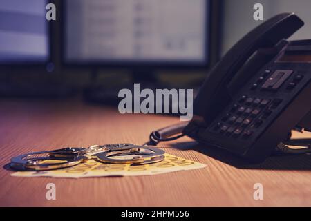 Festnetztelefon und Euro-Geld mit Handschellen auf dem Tisch, Nahaufnahme. Schreibtisch in einem dunklen Nachtraum mit Computermonitoren Stockfoto