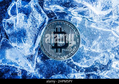 Foto einer blau getönten Bitcoin-Münze, die auf einer rissigen Eisfläche liegt. Stockfoto