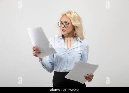 Seriöse Geschäftsfrau in formeller Kleidung und Brille beim Lesen von Dokumenten über hellgrauem Studiohintergrund Stockfoto