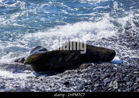 Gezeiten brechen über einem ungewöhnlich geformten Felsen an einem steinigen Strand in der Nähe von Playa de San Juan, Teneriffa, Kanarische Inseln, Spanien Stockfoto