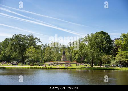 Menschen, die sich im Gras an der Statue von Joost van den Vondel im Vondelpark in Amsterdam, Niederlande, entspannen und sonnen Stockfoto