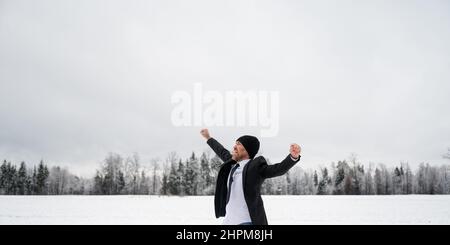 Begeisterter junger Geschäftsmann mit einem großen Lächeln auf dem Gesicht, der seinen Erfolg draußen in einer weißen, schneebedeckten Winterlandschaft feiert. Stockfoto