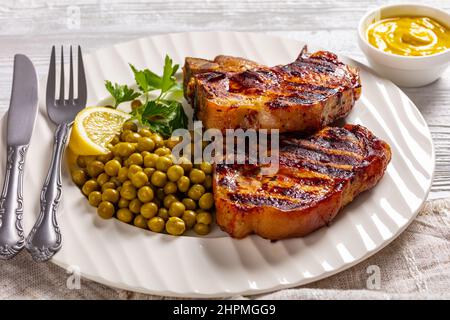 Gegrillte Schweinefleisch-T-Bone-Steaks mit gekochten grünen Erbsen und Zitrone auf einem weißen Teller auf einem Holztisch mit Senf, horizontale Ansicht Stockfoto