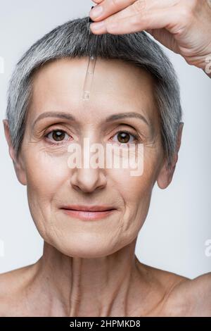 Nahaufnahme einer Frau mittleren Alters, die Vitamin C-Serum isoliert auf Grau anwendet Stockfoto