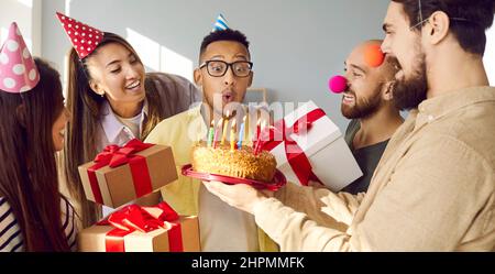 Fröhlicher junger Mann, umgeben von Freunden, die Kerzen auf den Geburtstagskuchen blasen, nachdem er sich gewünscht hat. Stockfoto