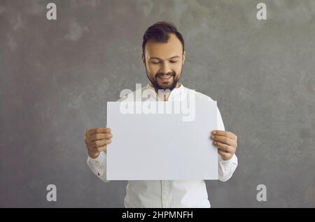 Glücklicher junger Kaukasusmann, der ein leeres Blatt Papier auf grauem Hintergrund hält. Stockfoto