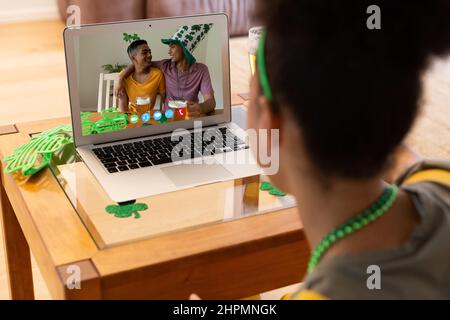 Frau mit gemischtem Rennen, die zu Hause mit zwei glücklichen männlichen Freunden einen Videoanruf zum St. patrick's Day auf einem Laptop führt Stockfoto