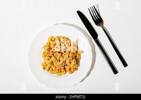 Lockige italienische Pasta mit Sauce auf einem weißen Teller. Minimalismus. Weißer Hintergrund. Pasta in Käsesauce. Messer und Gabel sind schwarz. Stockfoto