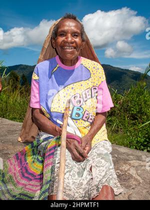 Alte Frau mit einem riesigen Lächeln in den Bergen von Papua-Neuguinea/PNG, die mit ihrem Gehstock und Bilum auf einem Felsen sitzt. Stockfoto