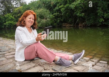 Reife hispanische Frau liest ein eBook auf einem elektronischen Leser, der an einem See in einem Park sitzt. Konzepte: Technik, Lesen und Entspannen im Freien. Horiz