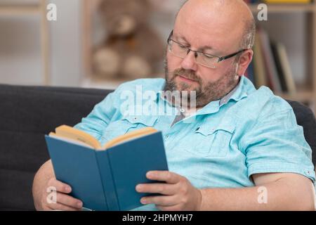 Ein kahler Mann, der auf einem Sofa sitzt, liest zu Hause ein Buch. Stockfoto
