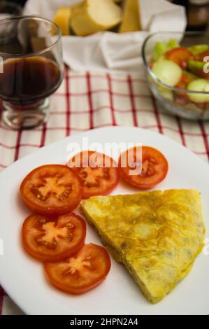 Teller mit einer Portion Kartoffelomelett mit Tomatenscheiben auf einem Tisch mit einem Glas Wein, Brot und einem Salat Stockfoto