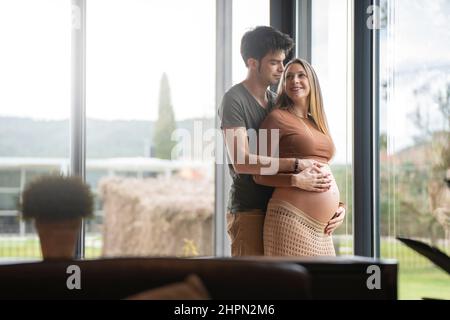 Die junge blonde Schwangere schaut aus dem Fenster und umarmt ihren Mann Stockfoto
