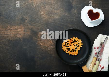 Herzförmige Spitzenpfannkuchen oder Crepes auf gusseiserner Pfanne auf altem rustikalem Holzhintergrund. Tischeinstellung für Ihr Lieblingsfrühstück am Valentinstag Stockfoto