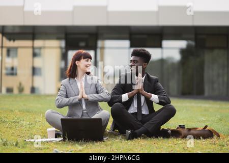 Porträt von ruhigen, entspannten jungen, multirassischen Bürokollegen, die draußen auf grünem Gras sitzen und Yoga-Meditation in namastiger Pose mit geschlossenen Augen machen. Büro Yoga und gedankenlos Stockfoto