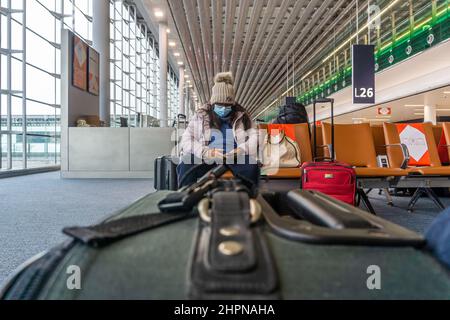 Flughafen Charles de Gaulle, Paris - eine weibliche ethnische Passagierin mit Gesichtsmaske und Checks auf ihrem Telefon, mit einem Koffer im Vordergrund Stockfoto