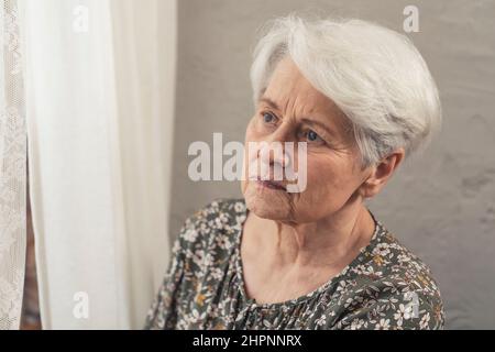 Wütend aufgeregt kaukasische ältere Frau durch das Fenster mit einem weißen Vorhang schauen. Hochwertige Fotos Stockfoto