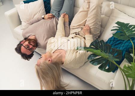 Glückliches Paar in ihren 30s auf dem Sofa in ihrer brandneuen Wohnung liegend, die Beine hochhebend und scherzend. Hochwertige Fotos Stockfoto