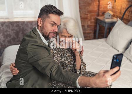 Ältere Großmutter und ihr ältester Enkel machen dumme Gesichter und machen ein Selfie mit schwarzem Smartphone. Hochwertige Fotos Stockfoto