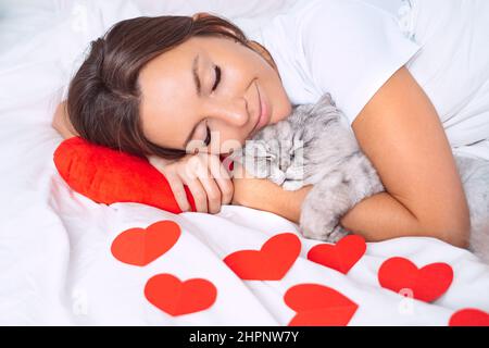 Schöne Frau, die mit roten Herzen auf dem Bett liegt und ihre schöne Katze umarmt. Liebe für Haustiere. Stockfoto