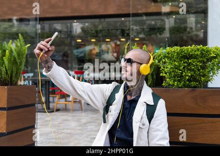 Mann hört Musik mit seinen Kopfhörern und nimmt ein Selfie auf einem Bürgersteig in der Stadt auf Stockfoto