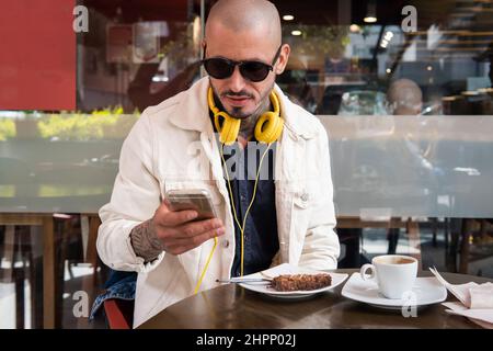 Ein junger Mann mit gelben Kopfhörern plaudert in einem Café mit seinem Handy, trinkt Kaffee und isst einen Kuchen Stockfoto