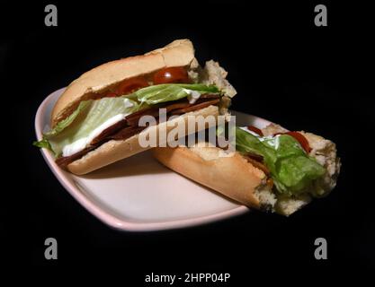 Vegan BLT - Eine Brotrolle mit veganem Speck, veganer Mayonaise, Salat und Tomaten im Inneren - auf einem herzförmigen Teller mit schwarzem Backgroun Stockfoto