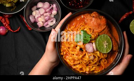 Draufsicht eine Frau, die Khao Soi Kai über schwarzem Tischhintergrund serviert. Nordthailändisches Essen im Restaurantkonzept. Nudeln, eine Pudelsuppe mit Hühnchen Stockfoto