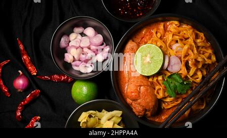Khao Soi Kai in einer schwarzen Schüssel serviert mit Beilage, Schalotten, eingelegten Senfgrüns, Limette und gemahlenen Chilis in Öl gebraten. Nordthailändisches Essen concep Stockfoto