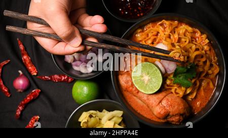 Eine weibliche Hände mit Essstäbchen essen Khao Soi Kai, cremig würzigen gelben Kurrinudeln Suppe mit Huhn serviert mit Beilage. Draufsicht Stockfoto