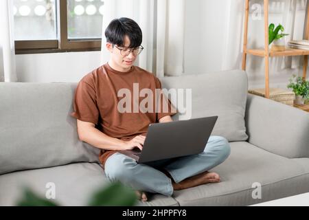Technologiekonzept der Mann, der in legerer Kleidung auf dem Sofa sitzt, sein Notebook auf dem Schoß auslaht und seine Arbeit verrichten will. Stockfoto