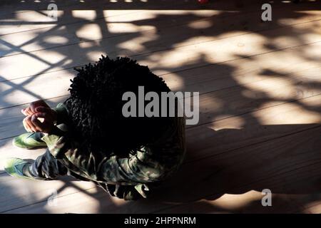 Allein von oben Vietnamesin sitzt drinnen auf Holzboden mit Schlagschatten von der Tür am sonnigen Nachmittag, die Menschen tragen Soldatenkleidung und zerzauste Haare Stockfoto