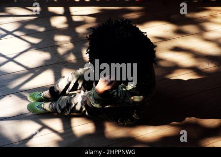 Allein von oben Vietnamesin sitzt drinnen auf Holzboden mit Schlagschatten von der Tür am sonnigen Nachmittag, die Menschen tragen Soldatenkleidung und zerzauste Haare Stockfoto