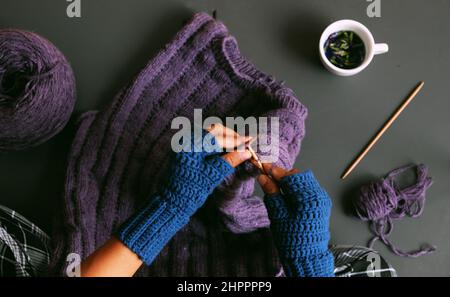 Draufsicht Frau Hand tragen blauen Fäustling Häkeln, stricken violetten Pullover als Geschenk im Winter, nahe Hand drücken Smartphone, um Foto auf dunklen Ton zu nehmen Stockfoto