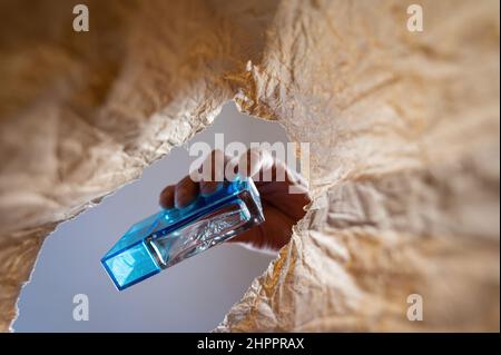 Eine Hand legt eine blaue Flasche Parfüm in einen Papierbeutel. Die Hand eines reifen Mannes hält die Glasflasche über dem offenen braunen Beutel. Von unten nach oben geschossen. Cl Stockfoto